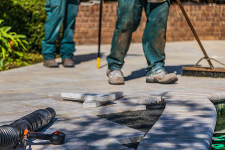 Workers laying patio tiles on a prepared base outdoors under sunlight during patio installation
