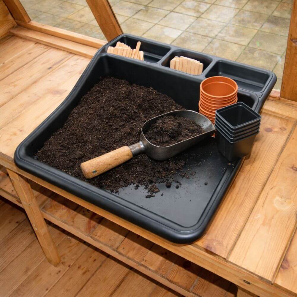 Black multi purpose garden tray being used for potting, holding a scoop of compost and small plant pots ready for filling and planting.