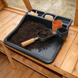 Black multi purpose garden tray being used for potting, holding a scoop of compost and small plant pots ready for filling and planting.